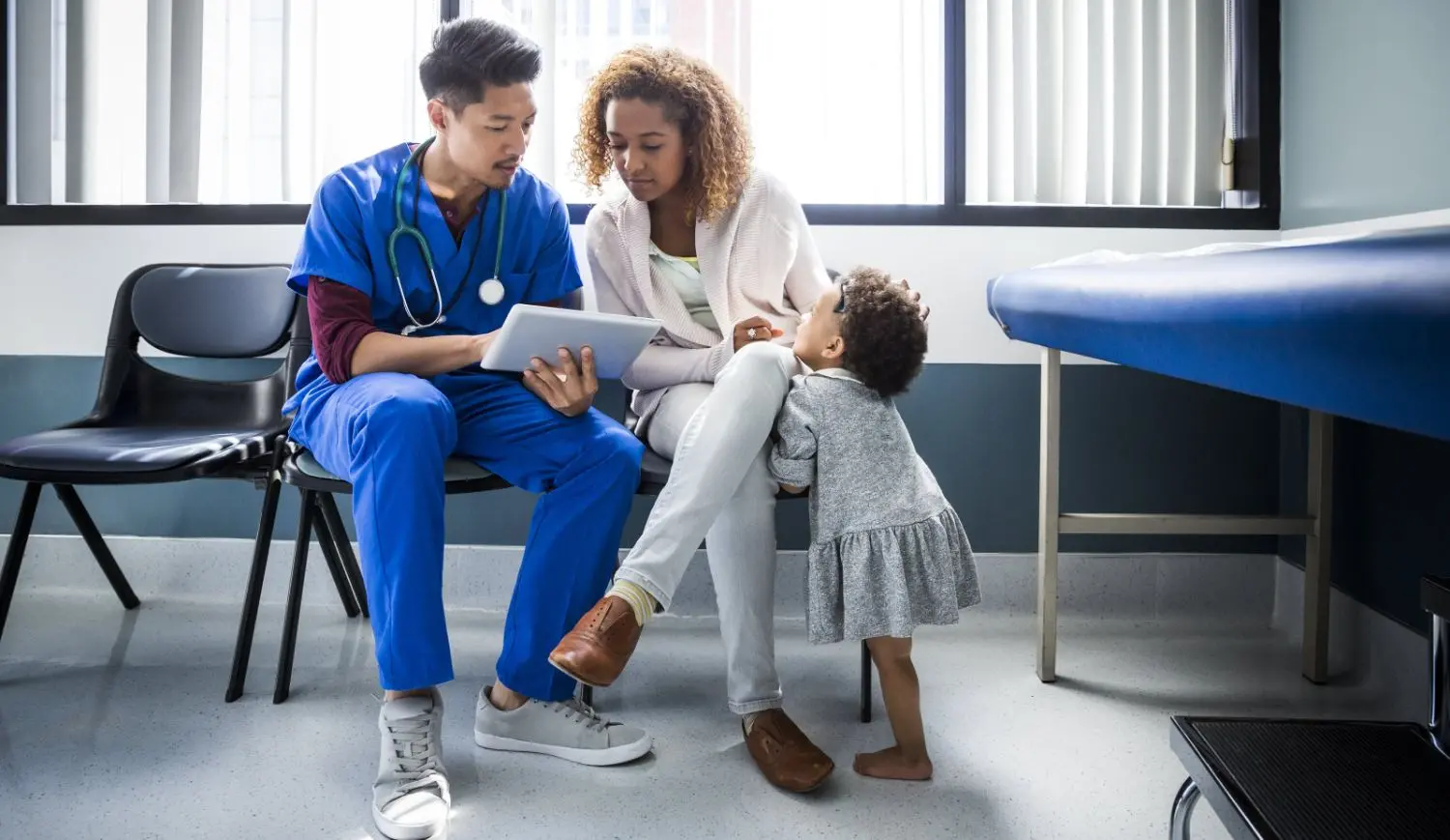 The picture is showing us possibly a consultation of a doctor and a mother with her daughter, they are sitting on chairs and doctor uses a tablet.