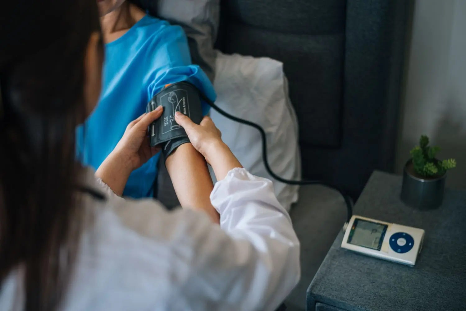 A woman sitting at a desk using a blood pressure monitor in a home setting with a plant visible in the background.