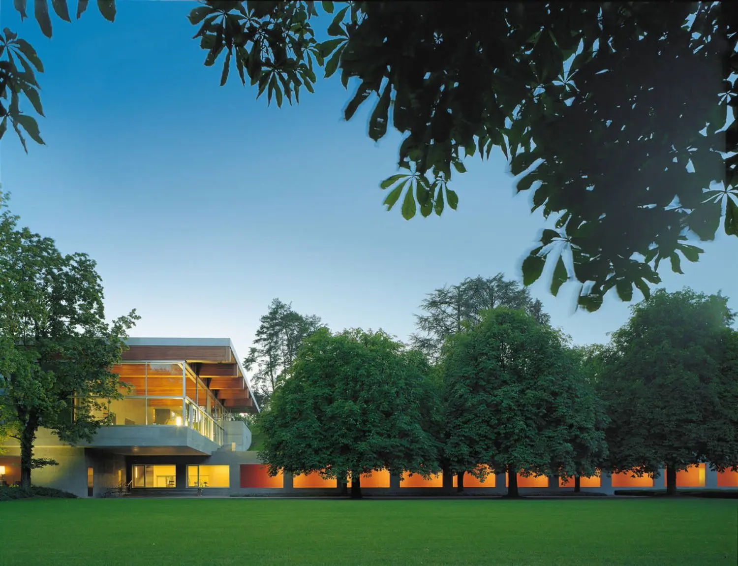 Wide shot of Swiss Re Centre for Global Dialogue featuring building and lit corridor with trees and grass in the foreground.
