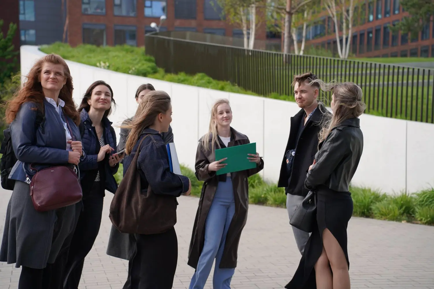A group of people are standing outside, engaged in conversation on a paved area with a modern building and greenery in the background.