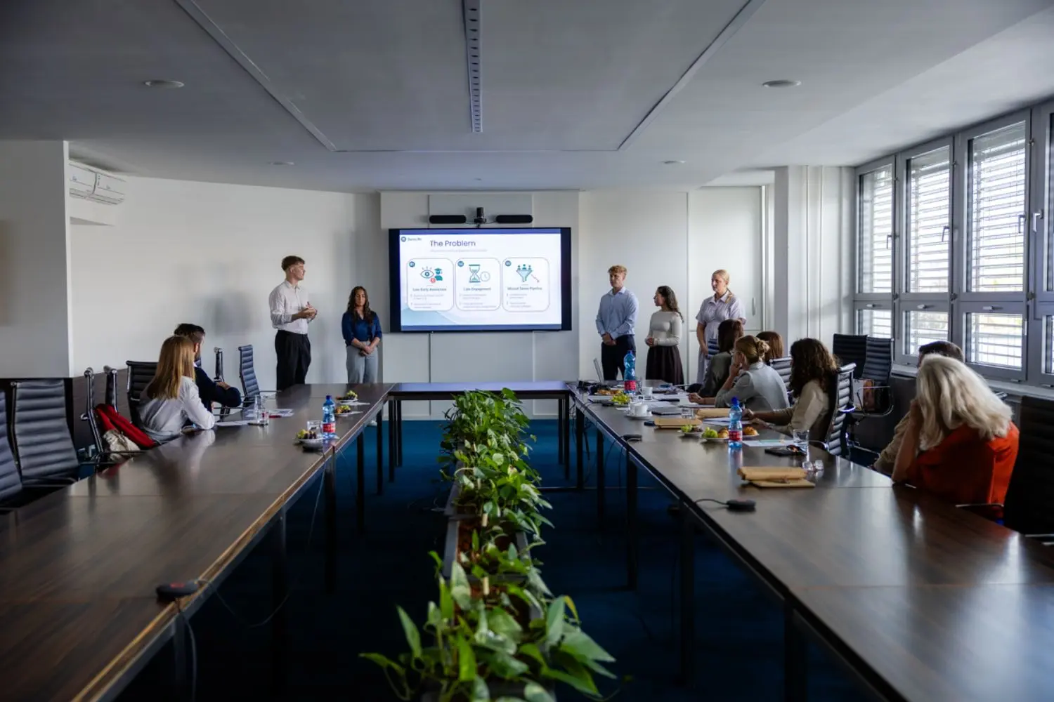 A conference room with a large table surrounded by people. A presentation is being displayed on a screen at the front, and presenters are standing up and speaking. A row of potted plants decorates the table, and sunlight streams through the windows.