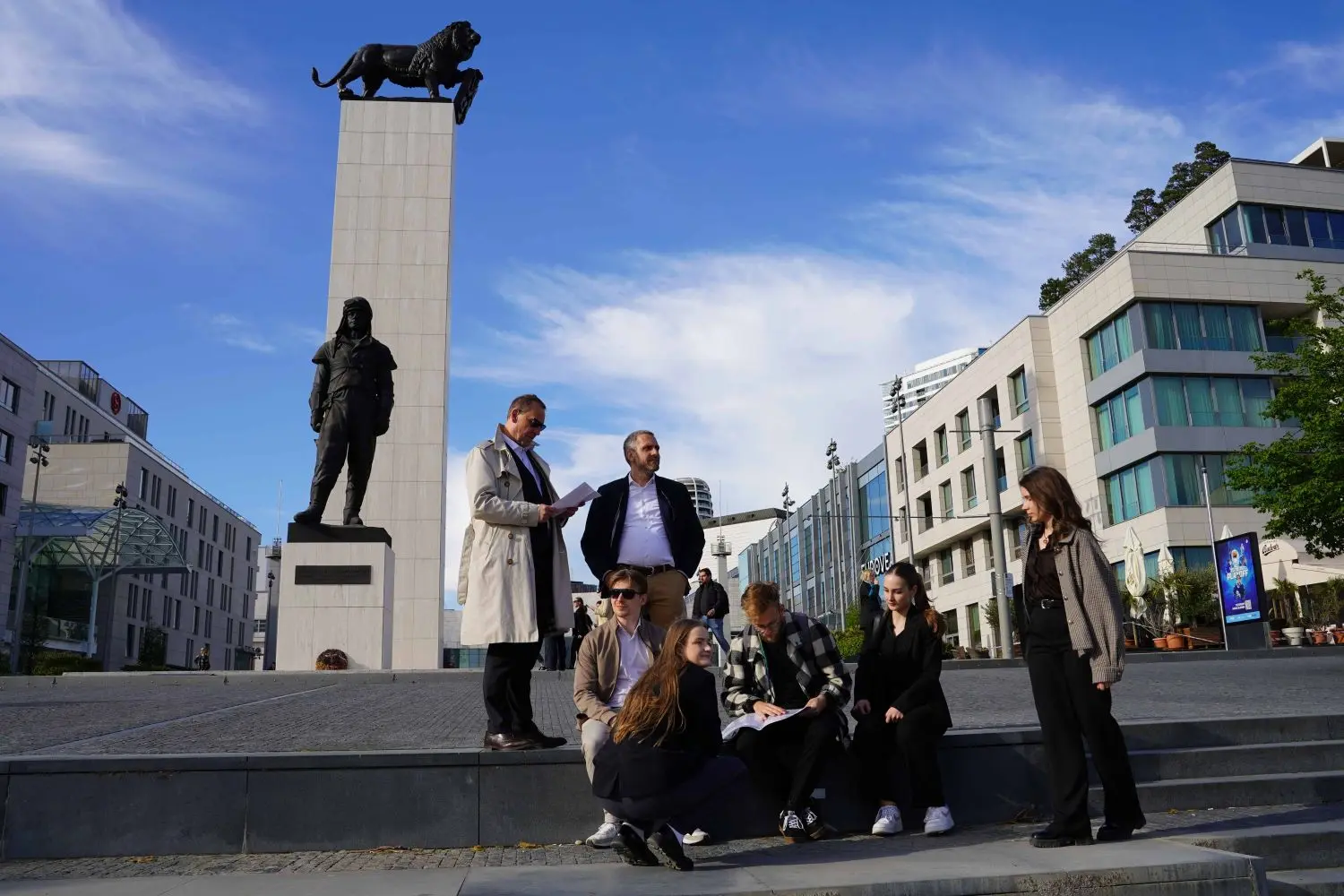 A group of people are gathered outdoors near a tall stone monument topped with a lion statue, with modern buildings in the background.