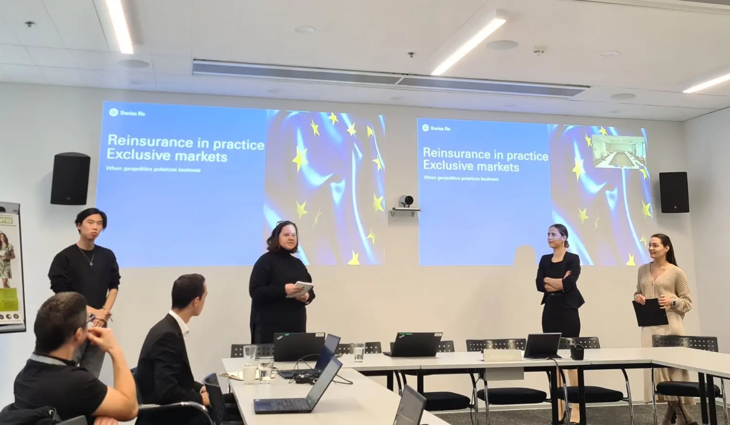 A presentation is taking place in a modern meeting room with multiple people standing and seated. Two large screens display the presentation topic about inclusion.